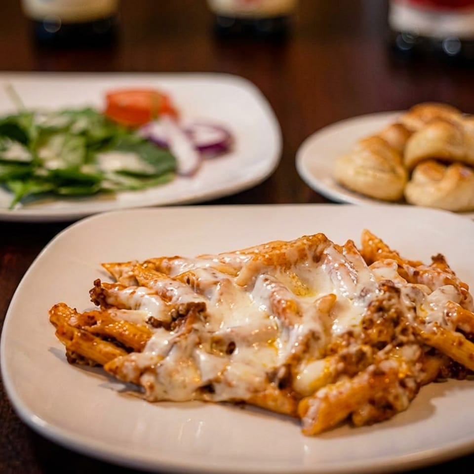 Baked penne pasta with melted cheese, a side salad, and bread rolls on a dark table.