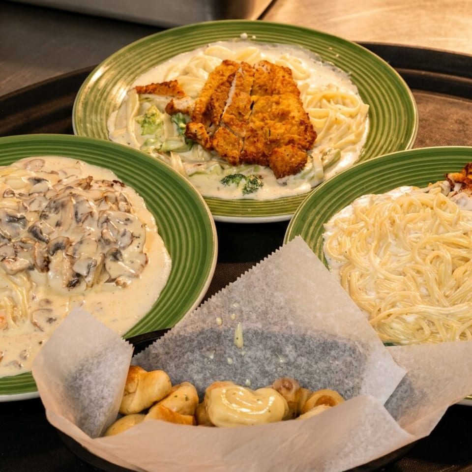Three green plates of creamy pasta with shrimp, chicken, and mushrooms, plus a basket of bread.